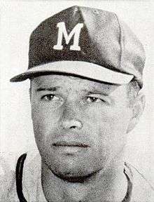 Bust portrait of a man in a white jersey with a dark stripe around the neck. He is wearing a dark baseball cap with a white "M" on it.