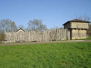 A wooden stockade fronted by a wall of vertically-placed logs and a corner blockhouse