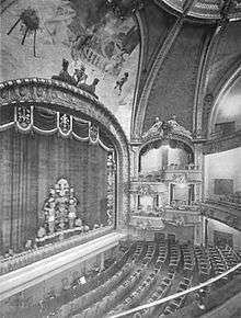 Black and white photo of the inside of a theater, with a high vaulted ceiling and an ornate curtain in front of the stage.