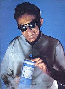 A picture of a blind man wearing dark glasses and a white coat grinning while holding a bottle of soap. Steam, presumably dry ice, billows from below, giving the photograph a science-fiction feel.