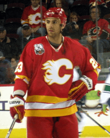 A hockey player stands and looks to his right during a pre-game warm up. He is wearing a red uniform with white and yellow bands at the waist and elbows, and a stylized "C" logo on his chest.