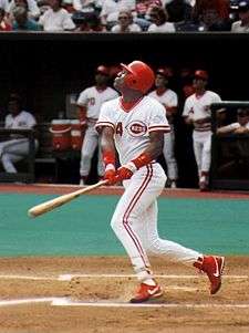 A dark-skinned young man wearing a white baseball uniform with red trim and a red batting helmet swinging a baseball bat