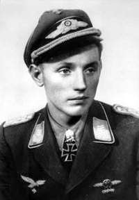 A black and white photograph of a smiling young man wearing a military uniform, peaked cap, various military decorations including a neck order in shape of an Iron Cross.