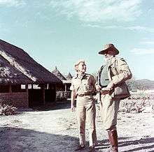  photograph of a man and woman with a brush covered hut in the background