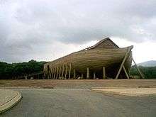 The fully constructed ark is at the center of the image with a downcast sky. A large ramp on the left side of the image is leading up into the ark. In the foreground of the image is a paved street.