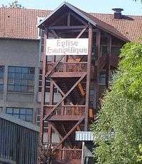 Wooden three-story chalet on stilts, with a white sign on the top floor reading "Église évangélique"
