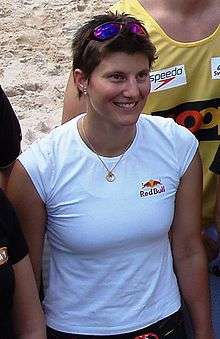 Young smiling woman with short brown hair, wearing a white T-shirt with the Red Bull logo and sleeves pulled back. She also wears a pair of sunglasses on top of her head and a golden chain with a circular pendant around her neck. She is on a beach among a group of people.