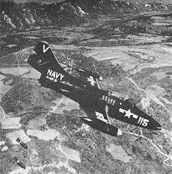 A single seat jet aircraft with US Navy markings in mid-air, flying in profile from left to right in a downward angle of attack below the camera. To the bottom left, two bombs are falling away from the aircraft towards an unseen target in the fields below.