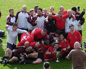 F.C. United players and coaches celebrate winning their first championship title. The championship trophy is seen on the green grass pitch and the group have their arms around one another.