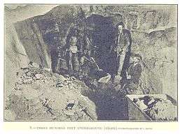 Inside a gold mine; men stand in a rough underground passage.