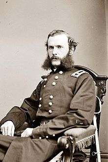 A white man with wavy hair and extremely bushy sideburns connected to a mustache, sitting in a chair. He is wearing a long double-breasted military coat with a rectangular patch over each shoulder.
