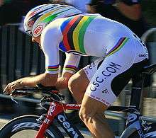 A road racing cyclist wearing a mostly white jersey with notable rainbow stripes across the midsection, biceps and hips, with a matching helmet, sitting crouched low on his bicycle.