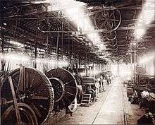 A photograph showing the interior of a large factory building with lighting provided by overhead skylights, underneath which is suspended a line shaft providing power via pulley belts to various machines on the factory floor, some of which dwarf workers standing by their stations