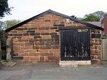 A low plain building in red sandstone with a gently sloping gabled roof and a door to the right with flaking black paint and a notice reading "Bridewell" in Gothic script