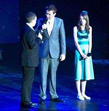 Three people on a stage, Johnston is being addressed by a man in a grey suit with a microphone. A teenage girl performer Faryl Smith, who has long brown hair and wears a green dress, is looking on.