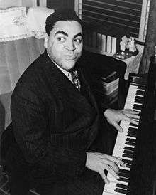 Stocky African-American man sitting and playing the piano. He has black hair and thick black eyebrows, and is grinning and looking to the left. The man is wearing a striped black suit, white shirt and a tie.