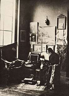 A seated man with a white beard reads a book at the corner of a studio.