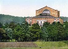 A building stands beyond a part-ploughed field and a row of trees. It has five sections. Farthest away, the tallest part with a v-shaped roof contains the stage. Adjoining it is the auditorium section built of patterned brick. Nearest is the royal entrance, made of stone and brick with arched windows and a portico. Two wings adjoin the auditorium.