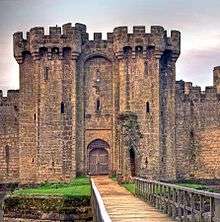 Photo of the main entrance which shows a large door recessed in a double archway in which there is a portcullis. The entrance is set in a tall battlemented gatehouse framed by twin towers with machicolations and slits. The entrance is approached by a bridge