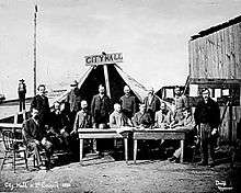 Men standing and sitting around two tables, facing the camera. A large tent behind them has a wooden sign that reads "City Hall"
