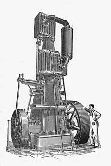A tall vertical steam engine with one small cylinder above a second larger cylinder. The engine is around 20 feet high and dwarfs the figure of a mechanic standing alongside.