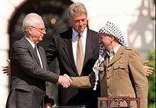 A man in a dark suit on the left shakes the hand of a man in traditional Arab headdress on the right. Another man stands with open arms in the center behind them.