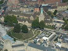 An aerial view of a town; a large grassy mound with two trees to the left; on the far side of the road, two adjoining rectangles of light-coloured stone two-storey buildings with dark tiled roofs, and a tall square stone tower topped with a short metal spire