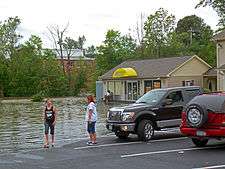 Two cars parked near the edge of water in a parking lot. A Subway restaurant can be seen in the background, and two young people are at the edge of the water.