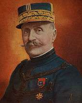 A gentleman with a handlebar-style moustache. He is wearing a peaked cap highly decorated with gold braiding. He wears a dark blue, high-collared jacket, with regularly spaced horizontal bands of dark braiding. On his left breast, he wears a medal attached to a red ribbon.