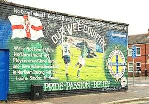 A large mural on a wall. Prominent features include the Ulster Banner, the IFA logo, the scoreline "Northern Ireland 1, England 0", and the captions "OUR WEE COUNTRY" and "PRIDE - PASSION - BELIEF"