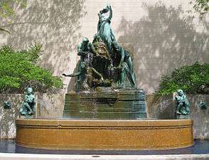 An outdoor fountain with water passing from one plate two another until it ends up in the fountain base.  The fountain has several women holding plates and is backed by a structural wall.  Trees are adjacent to the fountain on either side.