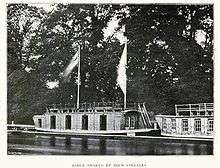 Two barges by the side of a river, one not fully in shot, in front of trees. The main barge has two large flags flying from the cabin roof, four large openings and one small one on the cabin's river side, and a window on the side of the cabin perpendicular to the river alongside a staircase to the roof; there are railings around the top of the roof. The other barge has five visible windows and roof railings.
