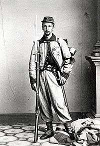 Full-body portrait of a white soldier with a rifle and bayonet by his side, standing on a crumpled flag. He is wearing baggy pants tucked into his boots, a short, decorative jacket buttoned at the top only, and a slouch cap.