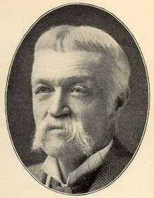 Head of an older white man with a drooping mustache wearing a checkered suit coat and tie.