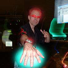 Stylized image of Frank Klepacki at his office wearing sunglasses and sticking his hand to the camera, red and green neon glow effects around his hair, hand, and sunglasses