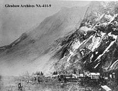 A small town shown at the base of a mountain.  The mountain's face stands barren following a large rockslide and a light cloud of dust is visible in the air.