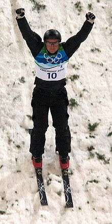 A man skis down a snow-covered slope with his arms in the air and fists closed. He is wearing black equipment with a blue-and-white vest bearing the Olympic rings and the number ten; a black helmet, black-and-white gloves, protective glasses, and red boots attached to black skis.