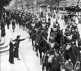 A long row of heavy cavalry stretches down a street, taking up most of the space. A woman in the foreground is reaching out and giving flowers to one of the men. They are wearing plate armour around the chest, and a crested hat on top.