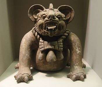 A fearsome mythical creature that may be either a bat or a jaguar. The head and face appear like that of a bat with a shortened snout, ridged eyebrows and very large round ears. Its mouth is open, showing pointed teeth and a protruding tongue. It wears a necklace made of two braided ropes, with an amulet in the front shaped the head of a double axe (or a bow tie). However, its body is not bat-like. It squats on four legs each with four clawed toes, with a perfectly round belly.