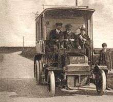 A sepia picture of an old bus, seen from the front. The cab is open to the elements and three men sit in it, while a boy stands to the right.