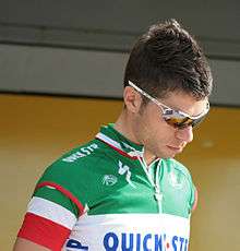 A man in his mid twenties wearing a cycling jersey bearing green, white, and red stripes. He wears sunglasses, and is looking downward.