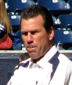 Color head-and-shoulders photograph of white man with dark (Gary Kubiak), wearing a white and navy sport shirt,  squinting in bright sunlight, with stadium seating in the background.