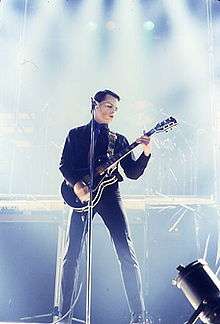 A colour photograph of Gary Numan performing onstage with a guitar and microphone