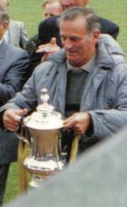 A middle aged man wearing shirt, grey pullover and light grey anorak, holding a trophy cup. other people behind him, all standing on grass. Bottom right of picture cut off by a wall.