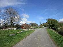 A sunny day in a green landscape, under a blue sky.  A country lane meanders from the middle foreground to traffic lights in the right midground. On the right is a grass verge and a hedge.  On the left, a grass verge edged with large boulders.  Beyond that a wooden rail fence marks the edge of a smaller road, and beyond that a building with a ridged pantile roof and large ornate bargeboards. This is 'station house' the last remnant of the railway hereabouts.