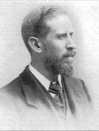 A black-and-white portrait photograph of a bearded man in a dark three-piece suit.