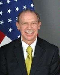 Full frontal photo of a white man's face and torso, smiling, with white mustache, blondish receding hair, slightly prominent ears, yellow necktie in half windsor, white shirt, dark suit, in front of American flag.