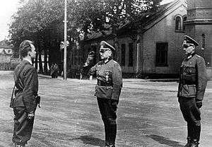 Three men standing, two on the left dressed in World War II German officer uniforms and saluting the man on the right