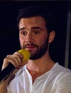 A 35-year old, dark-haired, bearded man, talking into a microphone and looking to the left of the camera.