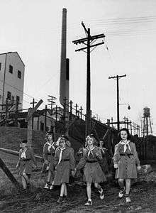 Five girls scouts in uniform. Two adult women in scout uniform watch over them. Behind them is a barbed wire fence, and in the background is an industrial building with a tall smoke stack.